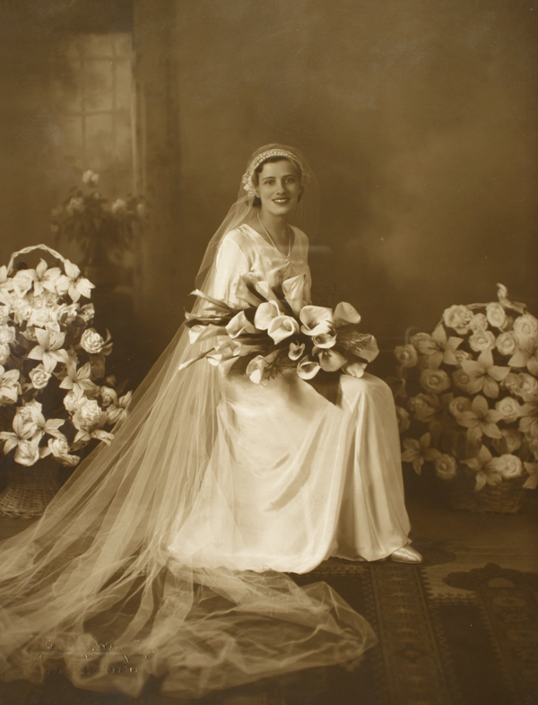 [Mujer vestida de novia con dos ramos grandes de flores a su alrededor y un ramo pequeño en sus manos]