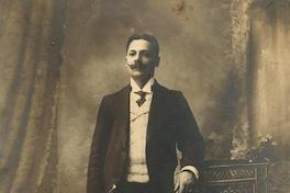 [Retrato de un hombre joven, pelo corto dividido al medio y bigote imperial; viste traje, chaleco, camisa cuello pajarita y corbata, posa de pie y apoya su mano izquierda en un sillón de mimbre]