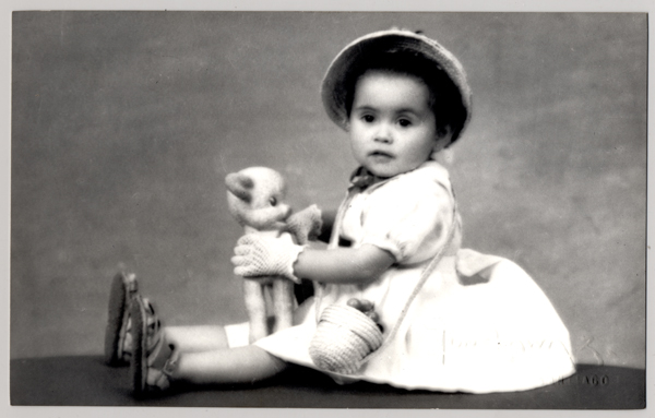 [Niñita sentada con un vestido blanco con sombrero y un canasto cruzado sobre sus hombros, ambos adornados con frutas y un juguete en sus manos]