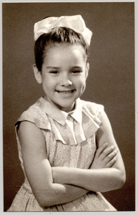 [Niña de unos ocho años, posando sonriente con un vestido de broderíblanco, con vuelos en el cuello, una cinta en el pelo y de brazos cruzados]