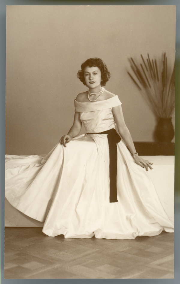 [Mujer posando con un vestido amplio y largo, hombros al desnudo, collar blanco de tres vueltas, sentada en una tarima y al fondo un macetero con espigas]