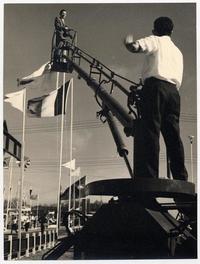 [Domingo Ulloa, con su cámara fotográfica, suspendido en el aire sobre una escalera mecánica para tener visión panorámica de la Exposición Ganadera de la Quinta Normal].