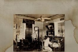 [Hombre sentado frente a su escritorio, de madera noble dividido en gavetas; alrededor de la habitación, cuadros, vitrina, anaquel con frascos de minerales, lámpara de colgar con brazos que sostienen tulipas de cristal; papel tapiz en sus muros]