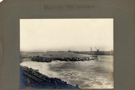 [Vista de unas vías férreas a la orilla del mar, de fondo se divisa el muelle]