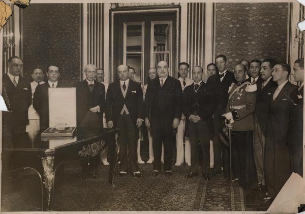 [Retrato de Manuel Rivas Vicuña, junto a un grupo de personas, en una ceremonia de condecoración; En una mesita, se encuentra la caja que guarda la distinción]