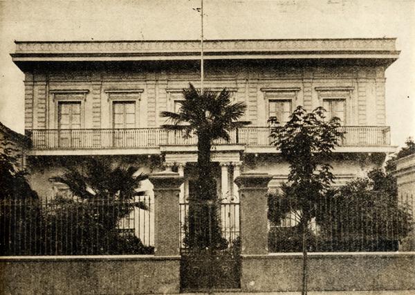 [Fachada de casa de dos plantas y un balcón con balaustres; la puerta de acceso está enmarcada por dos parejas de columnas; ante jardín y reja]