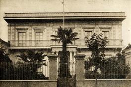 [Fachada de casa de dos plantas y un balcón con balaustres; la puerta de acceso está enmarcada por dos parejas de columnas; ante jardín y reja]