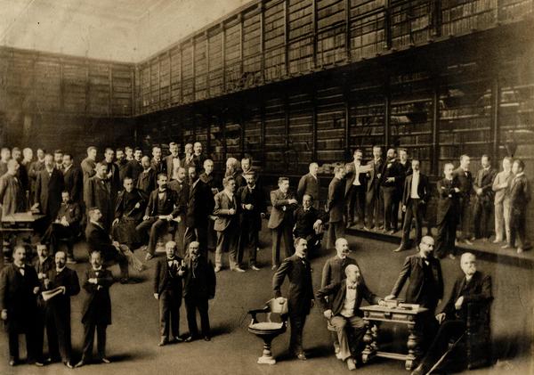 [Fotomontaje de un retrato colectivo: Un grupo de hombres, se encuentran al interior de la (biblioteca del Instituto Nacional), se observan mesas, estanterías y sillas, sentado a la extrema derecha, Diego Barros Arana]