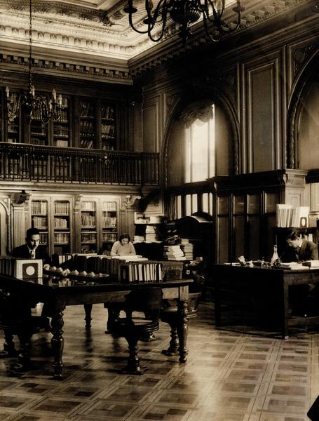 [Vista interior del fondo Bibliográfico Sala Medina, Biblioteca Nacional: Espacio revestido en madera de nogal, Anaqueles con puerta de vidrio, parquet de madera noble, lámparas de techo en bronce, de 8 brazos con bombillas como velas, balconada en el segundo piso, mobiliario de maderas nobles]