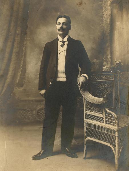 [Retrato de un hombre joven, pelo corto dividido al medio y bigote imperial; viste traje, chaleco, camisa cuello pajarita y corbata, posa de pie y apoya su mano izquierda en un sillón de mimbre]