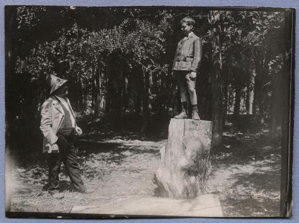 [En medio de un bosque, un niño posa sobre un tronco de árbol, un señor mayor observa]