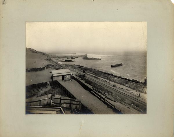 [Vista panorámica del puerto, se divisan vías férreas a orillas del mar, unas casas de la faena, y barcos en el mar]