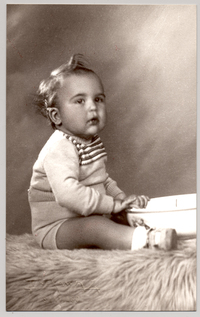 [Niñito rubio en traje de lana pantalón corto y chaleco con rayas sentado sobre un felpudo con un lavatorio entre sus piernas]