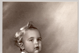 [Niñito rubio en traje de lana pantalón corto y chaleco con rayas sentado sobre un felpudo con un lavatorio entre sus piernas]