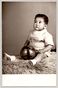 [Niñito de pantalón corto con cinturón sentado sobre un felpudo con una pelota y mirando hacia arriba]