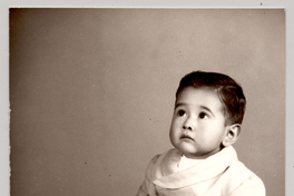 [Niñito de pantalón corto con cinturón sentado sobre un felpudo con una pelota y mirando hacia arriba]