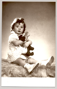 [Niñita de pelo rizado, vestido blanco abrazada a un juguete sentada sobre un felpudo]