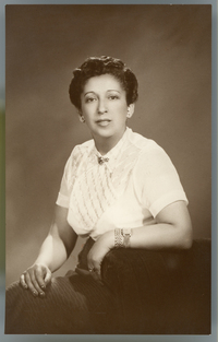 [Mujer morena de pelo corto, blusa blanca manga corta y un prendedor en el cuello, sentada en un sillón de estudio]