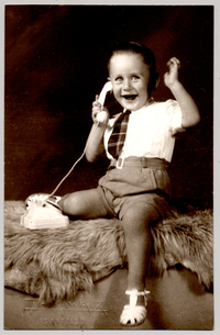 [Niño sonriente sentado sobre un felpudo con pantalones cortos, camisa blanca manga corta, corbatín a rayas y chalas blancas, simulando hablar por un teléfono de juguete]