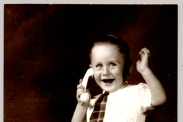 [Niño sonriente sentado sobre un felpudo con pantalones cortos, camisa blanca manga corta, corbatín a rayas y chalas blancas, simulando hablar por un teléfono de juguete]