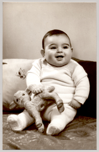 [Bebé moreno con un traje de lana color blanco sentado sobre una colchoneta de brocato con dos plumones en sus espaldas y un juguete en sus manos]