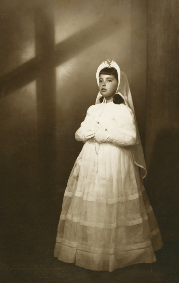 [Niña de Primera Comunióncon una amplia toca amarrada bajo el cuello y velo largo]