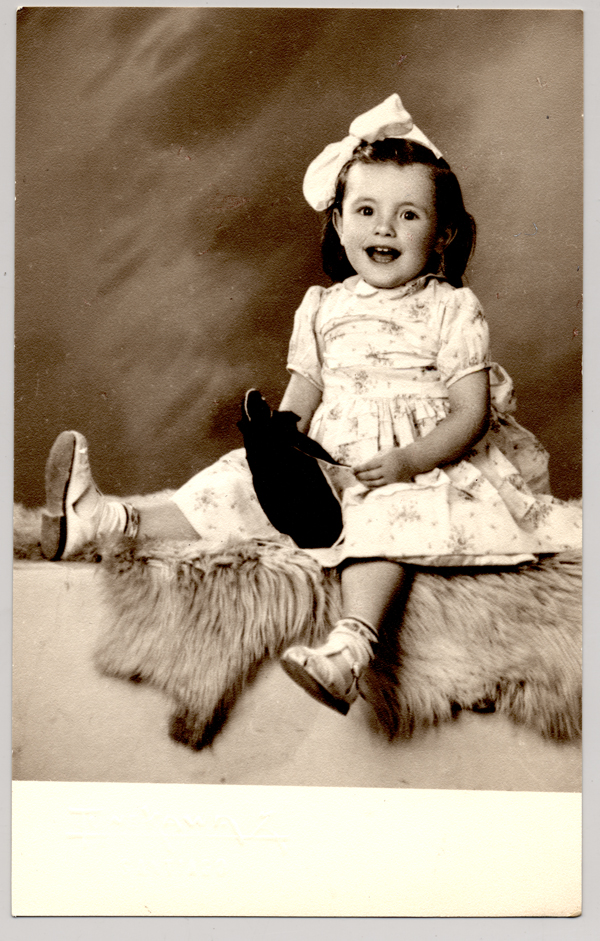 [Niñita sentada sobre un felpudo con un vestido repolludo con flores, sandalias blancas con soquetes, una cinta en el pelo y con un juguete en sus manos]