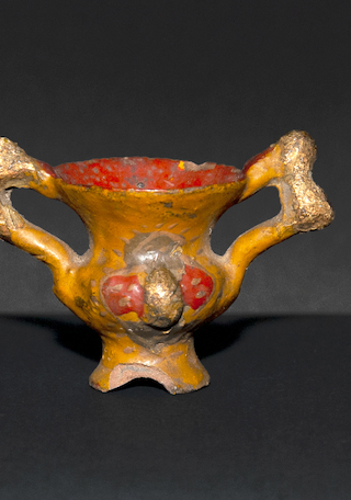 Vasija en miniatura con dos asas polilobuladas y doradas, con fragmentos faltantes.